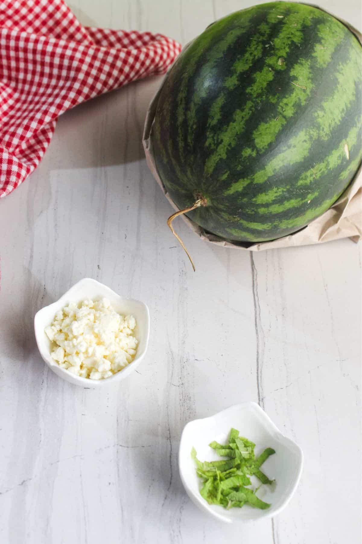 A whole watermelon, a small bowl of crumbled cheese, and a small bowl of chopped herbs sit on a white surface beside a red checkered cloth.