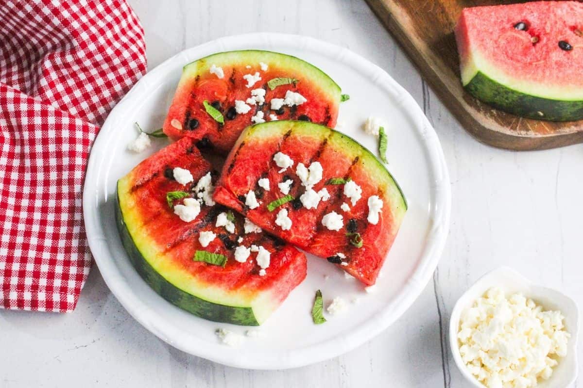 Three watermelon slices topped with crumbled cheese and mint are arranged on a white plate, with a red checkered cloth and extra watermelon and cheese nearby.