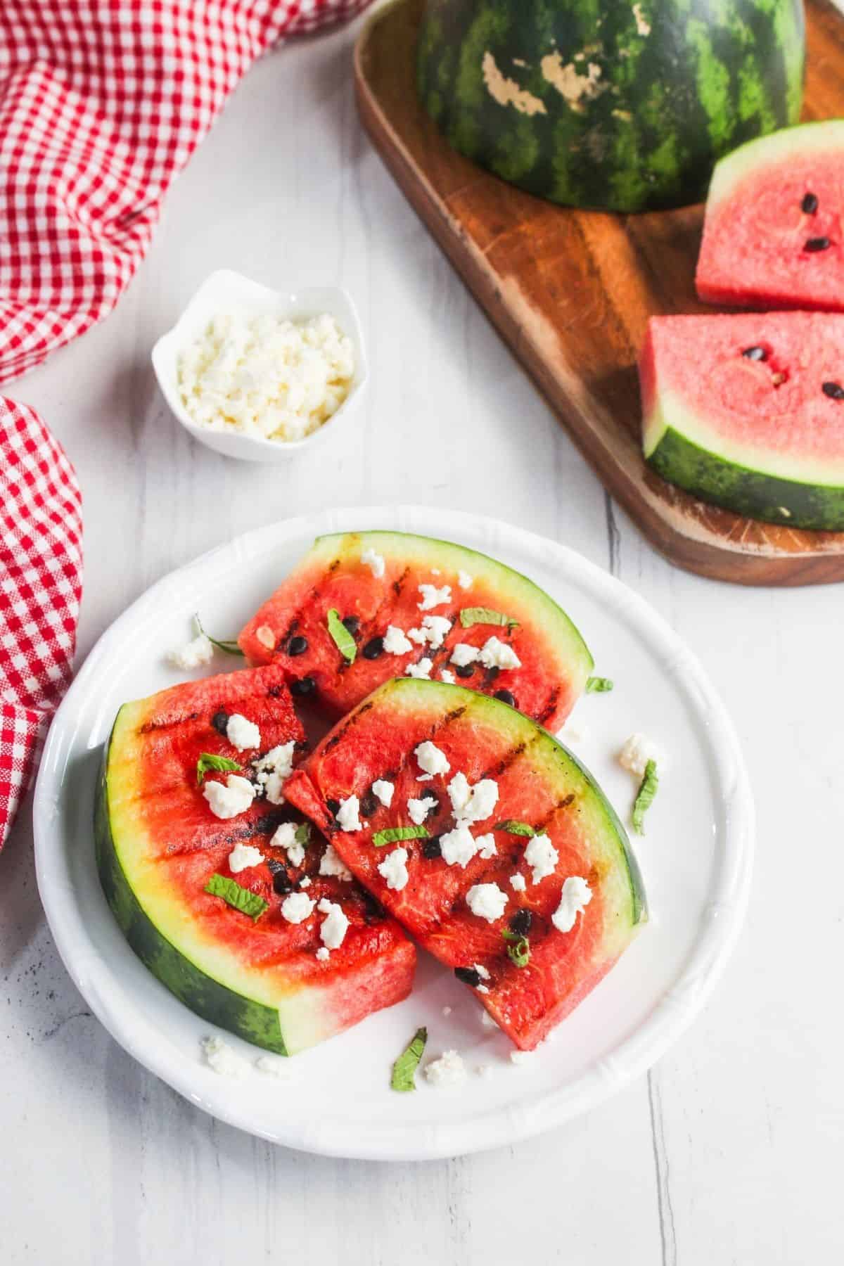 Sliced watermelon on a white plate topped with crumbled cheese and herbs, with extra cheese in a small dish and more watermelon on a cutting board in the background.