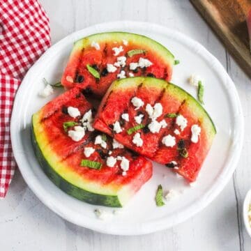Three grilled watermelon slices topped with crumbled cheese and mint are arranged on a white plate, next to a red checkered napkin.