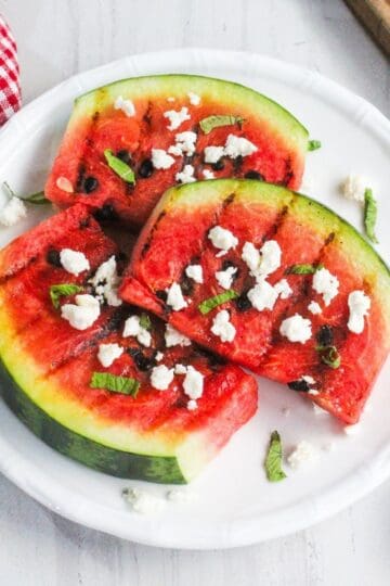 Three grilled watermelon slices topped with crumbled cheese and mint are arranged on a white plate, next to a red checkered napkin.