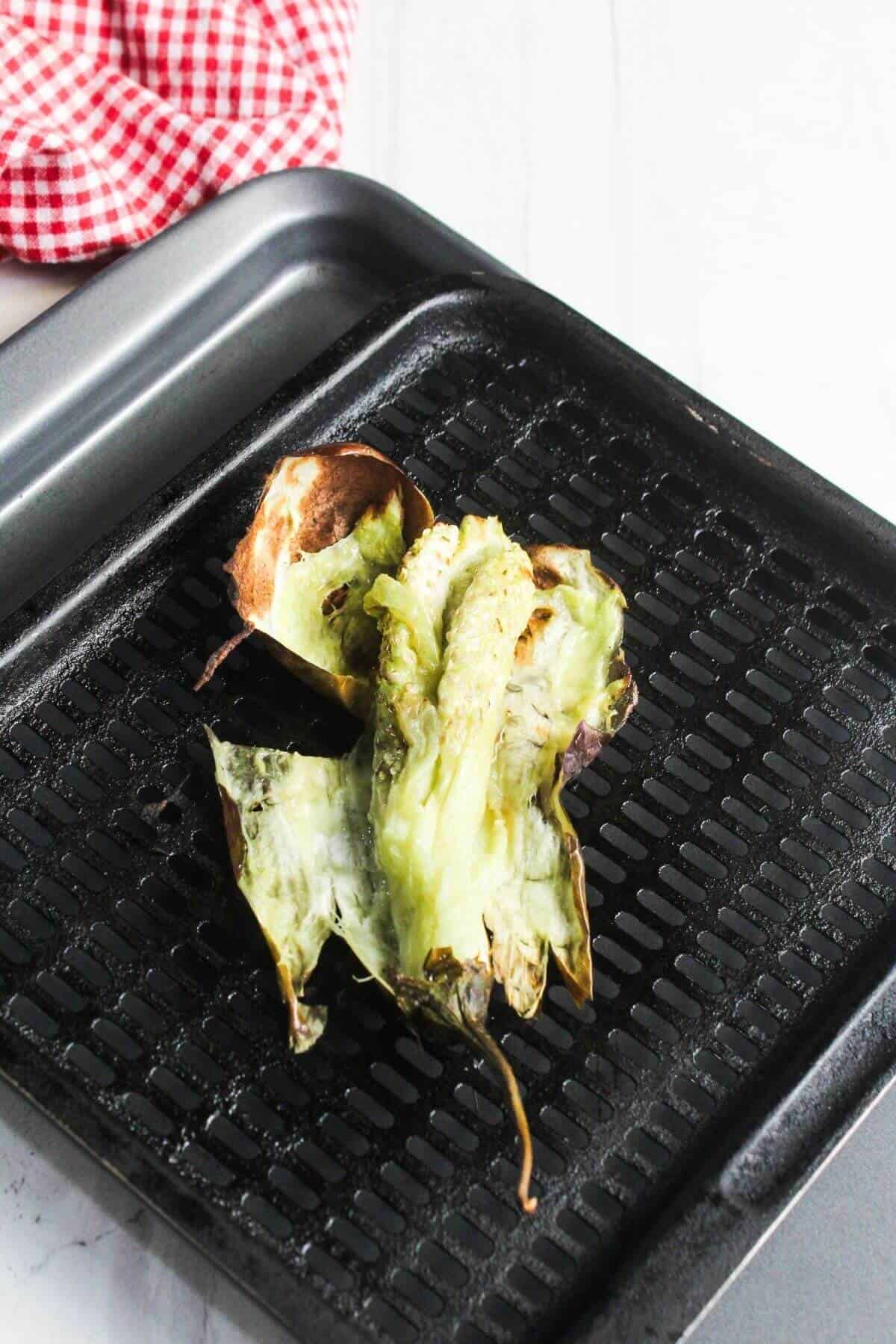 Charred and softened tomatillo husks and stems on a black grill pan, with a red and white checkered cloth in the background.