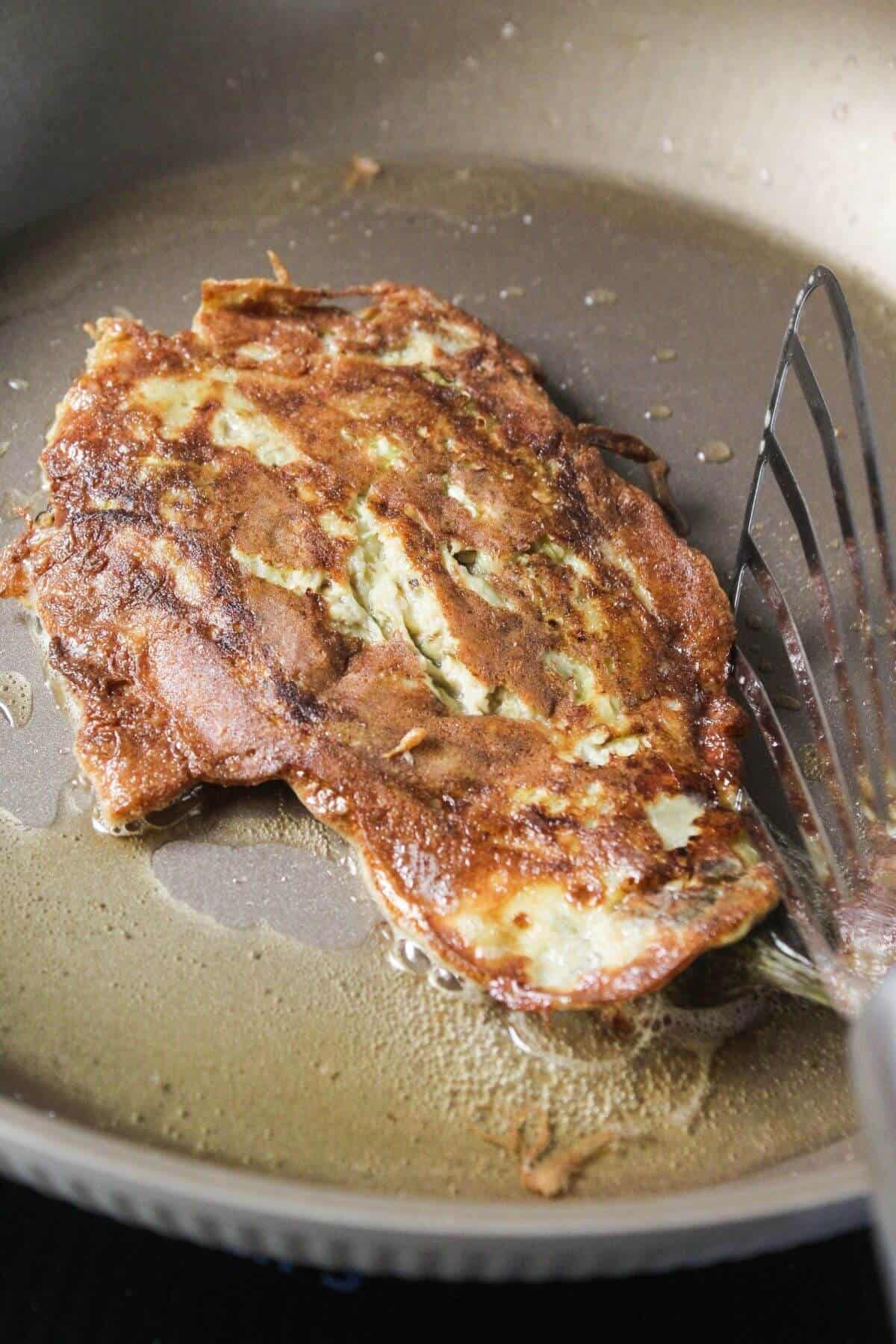 A browned omelette is being cooked in a frying pan, with a metal spatula positioned underneath it.