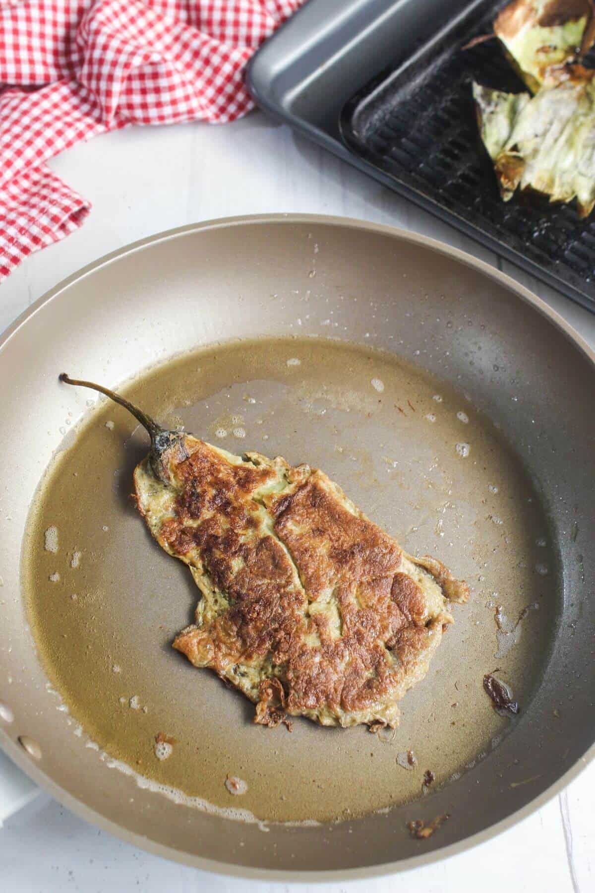 A cooked, battered chile relleno sits in a frying pan, with some oil visible. A baking tray and a red checkered cloth are in the background.