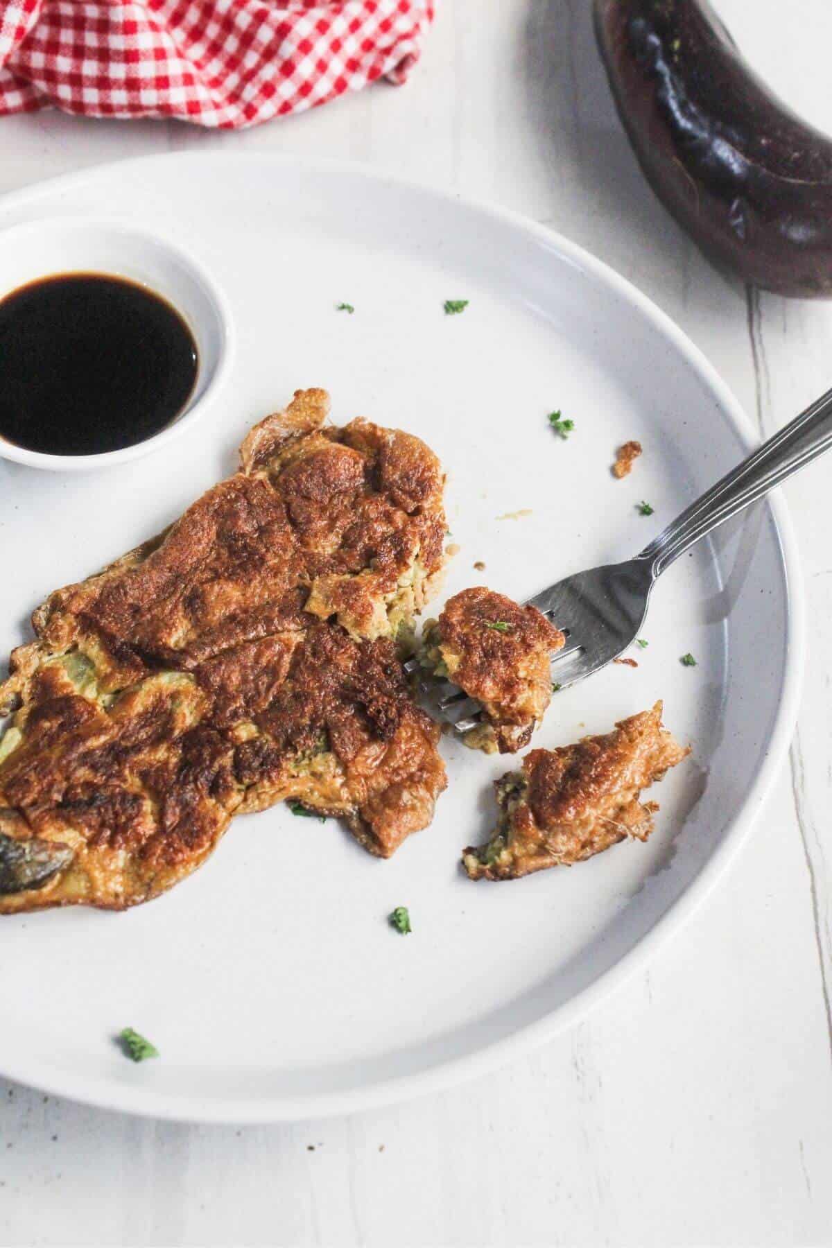 A piece of eggplant omelette is being cut with a fork on a white plate, with a small dish of dark sauce and a whole eggplant nearby.