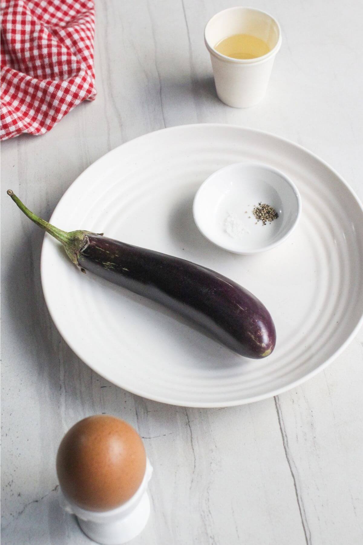 A whole eggplant on a white plate with a small bowl of salt and pepper, an egg in an egg cup, a paper cup of liquid, and a red checkered cloth on a white surface.