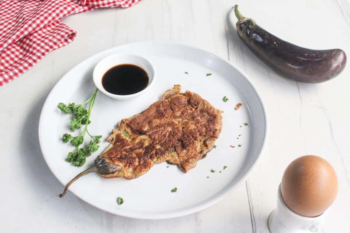 A cooked eggplant omelette on a white plate with soy sauce, parsley garnish, a whole eggplant, an egg, and a red checkered cloth on a white surface.