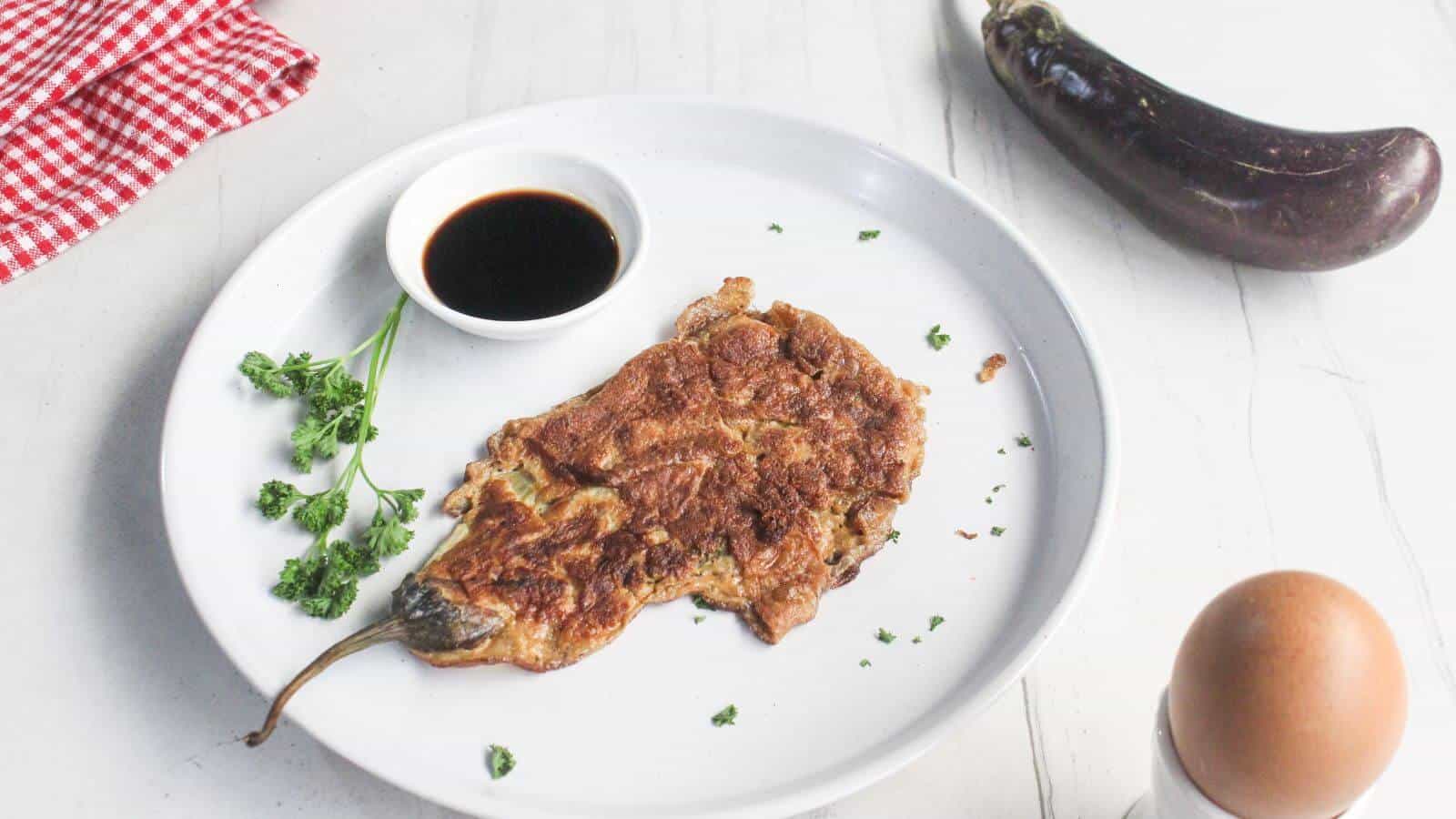 A cooked eggplant omelette on a white plate with parsley, a small dish of soy sauce, an egg, and a whole eggplant nearby.