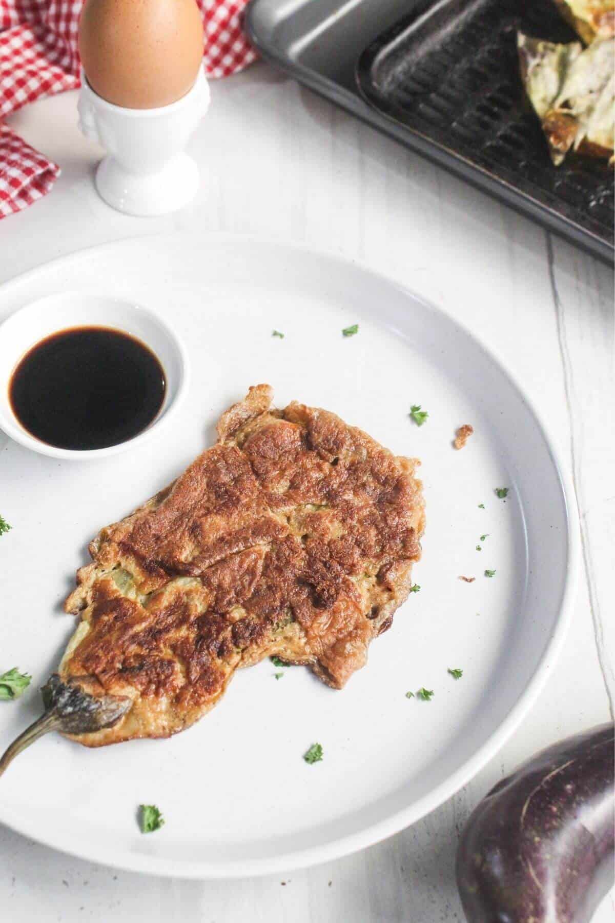 A cooked eggplant omelette on a white plate with a small dish of soy sauce, garnished with chopped herbs. An egg in an egg cup and a tray of food are in the background.