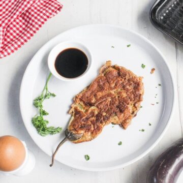 A plate with a cooked eggplant omelette, a small bowl of dark sauce, and a sprig of parsley on a white surface. An egg, a red checked cloth, and a grill pan are nearby.
