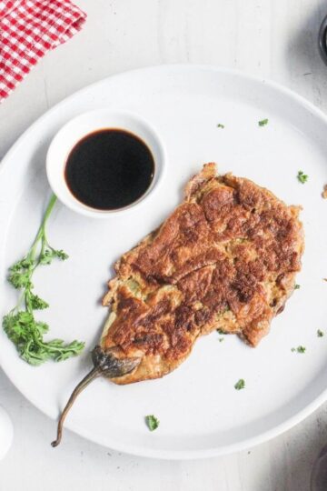 A plate with a cooked eggplant omelette, a small bowl of dark sauce, and a sprig of parsley on a white surface. An egg, a red checked cloth, and a grill pan are nearby.