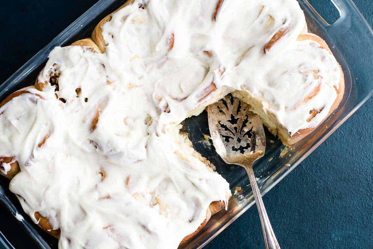 A glass baking dish with frosted cinnamon rolls, one missing piece, and a metal serving spatula resting in the dish.