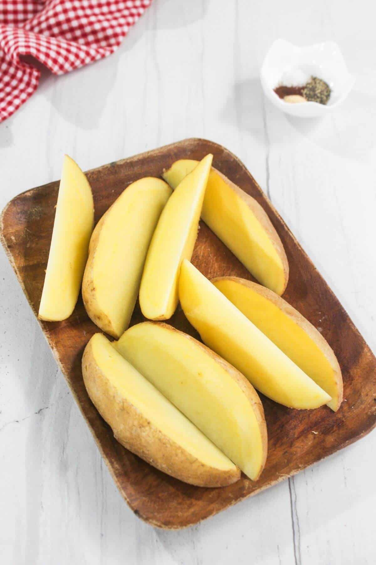 A wooden plate holds six raw potato wedges, placed on a white surface. A small bowl with spices and a red checked cloth are in the background.