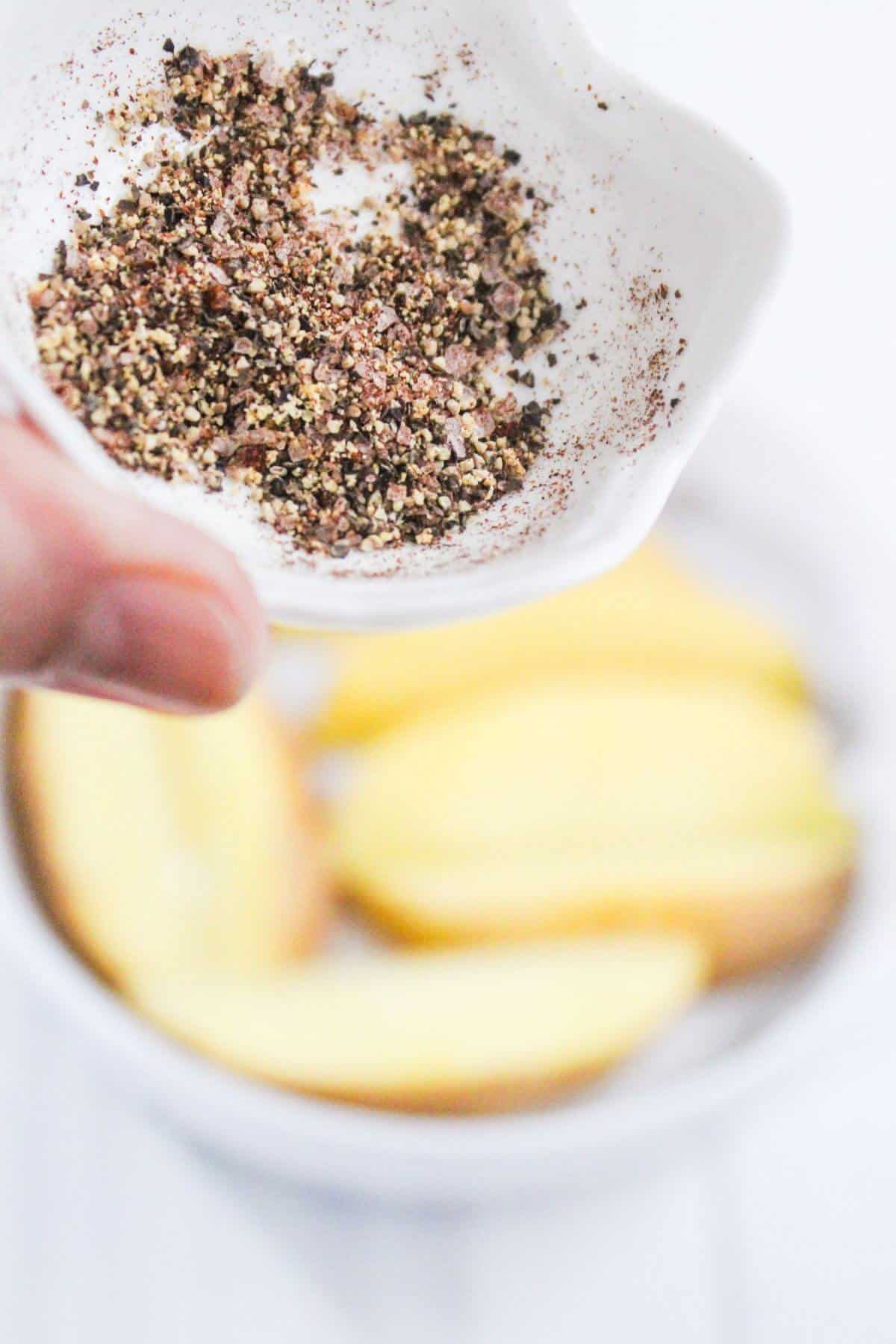 A hand holds a small white dish filled with ground black pepper above a bowl of sliced yellow apples.