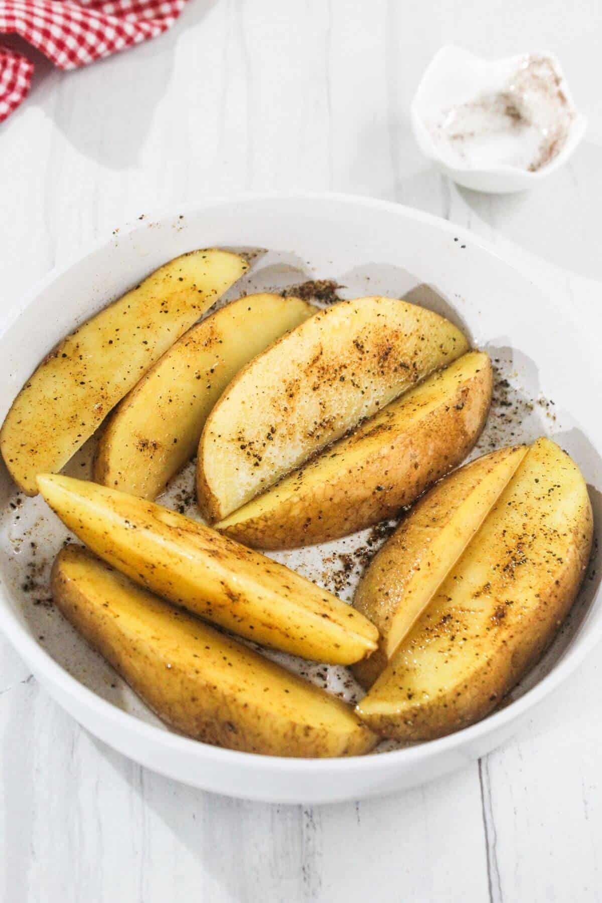 Potato wedges seasoned with spices in a white bowl, on a white surface. A small bowl of salt is in the background, along with a red and white checkered cloth.