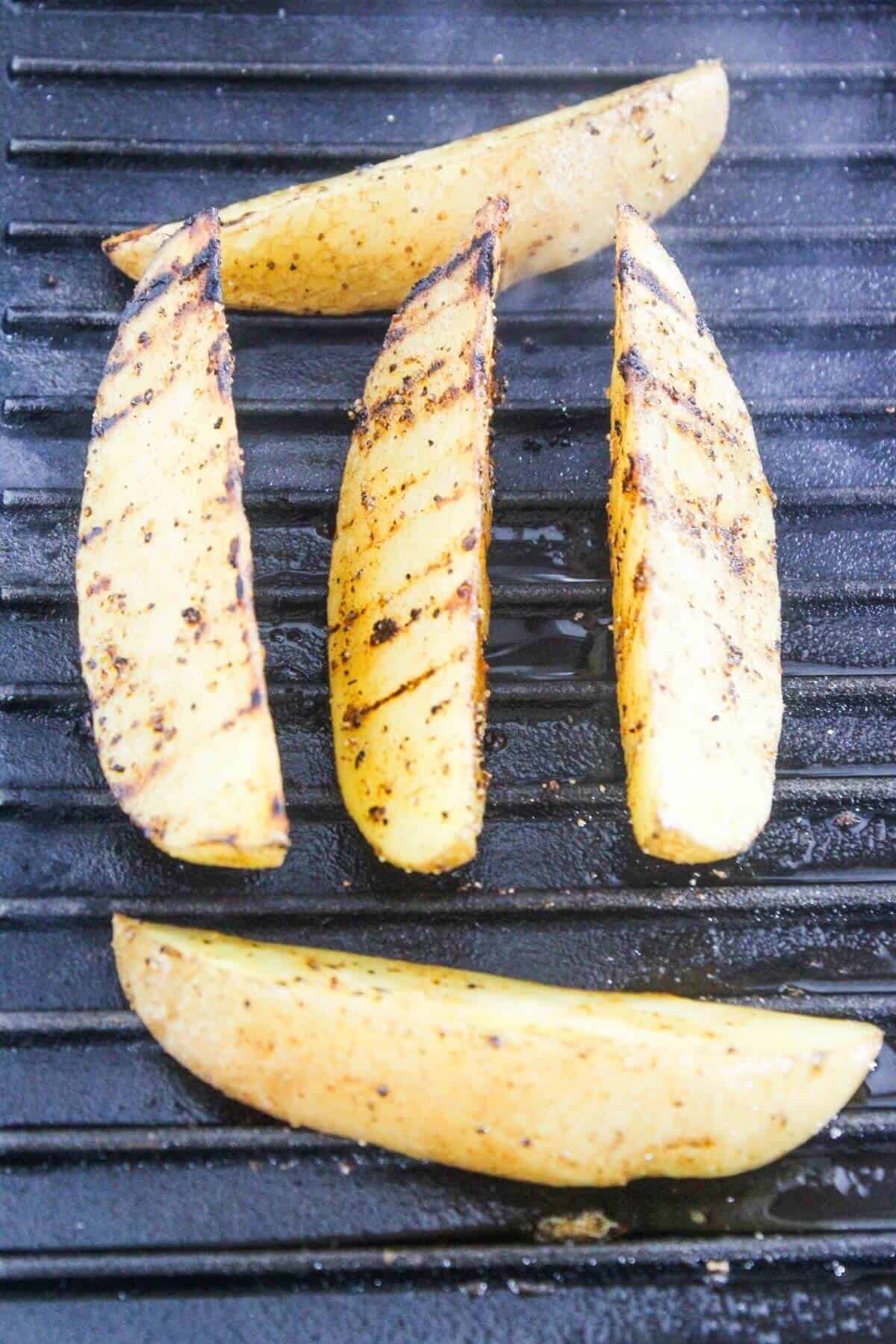 Four seasoned potato wedges with grill marks are cooking on a black grill grate.