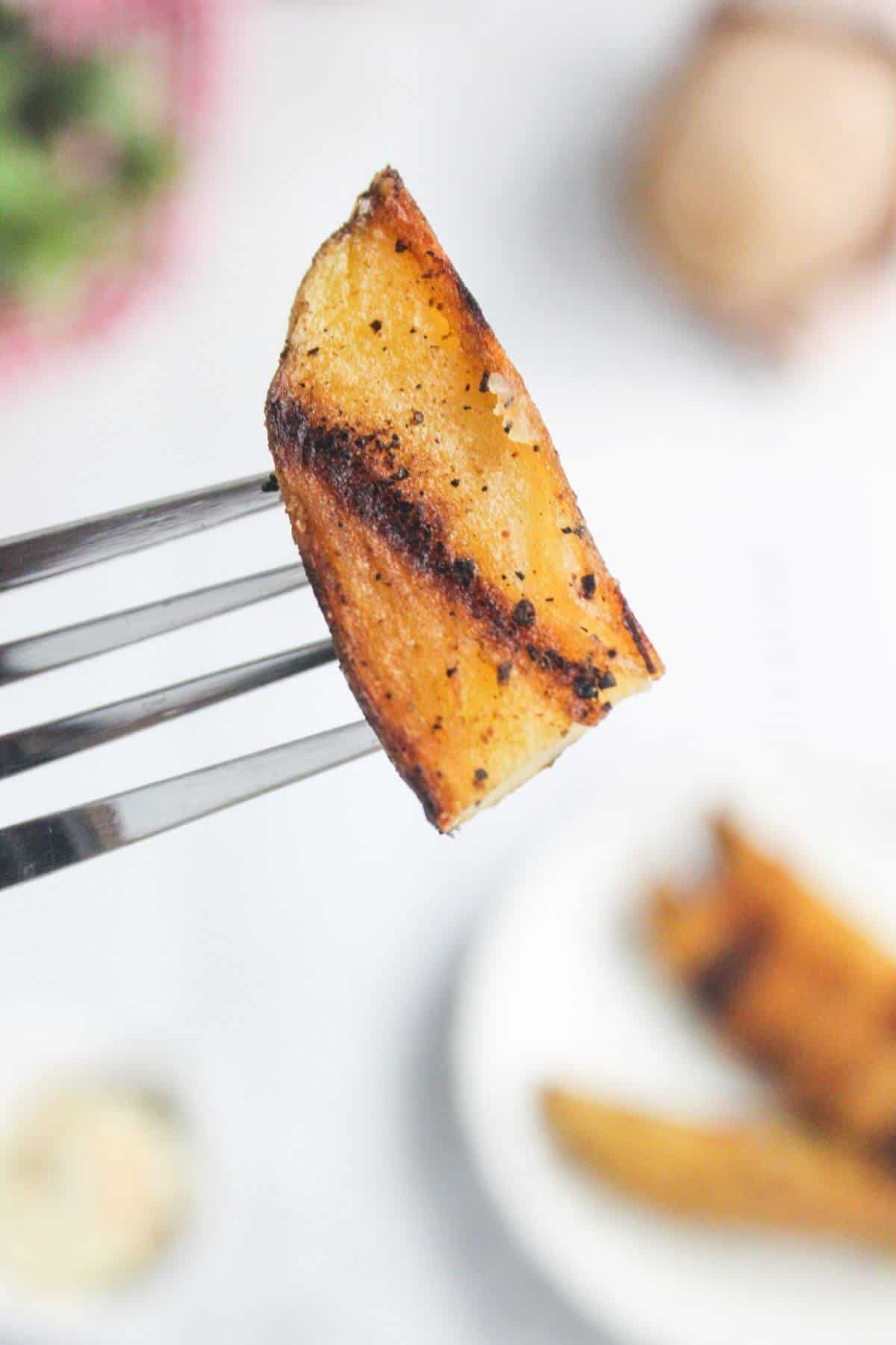 A fork holds a grilled potato wedge seasoned with spices, with more potato wedges and a blurred plate visible in the background.
