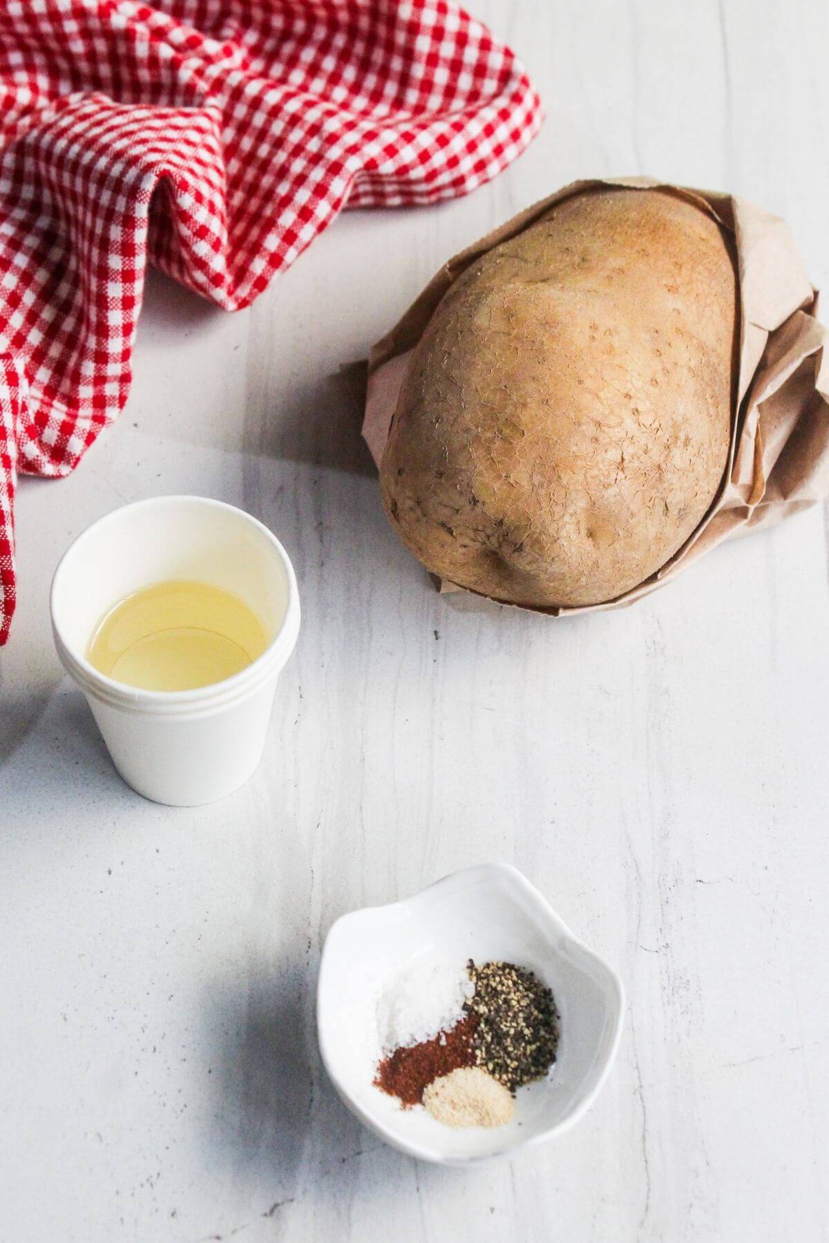 A wrapped vegan roast, a cup of oil, and a small dish with salt, pepper, garlic powder, and paprika are arranged on a white surface beside a red and white checkered cloth.