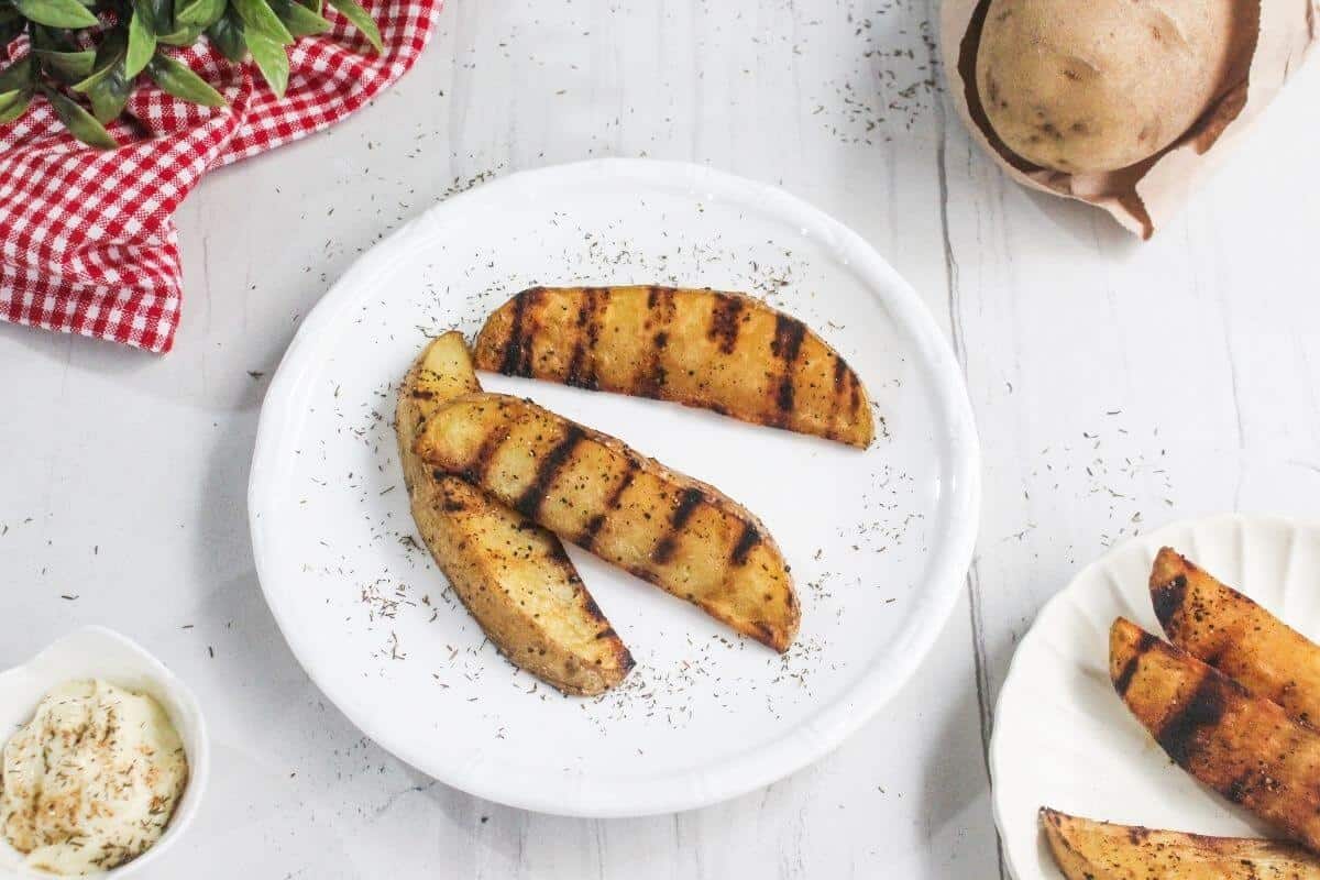 Three grilled potato wedges on a white plate, sprinkled with herbs. A whole potato, a dish of dip, and more wedges are nearby on a white surface.