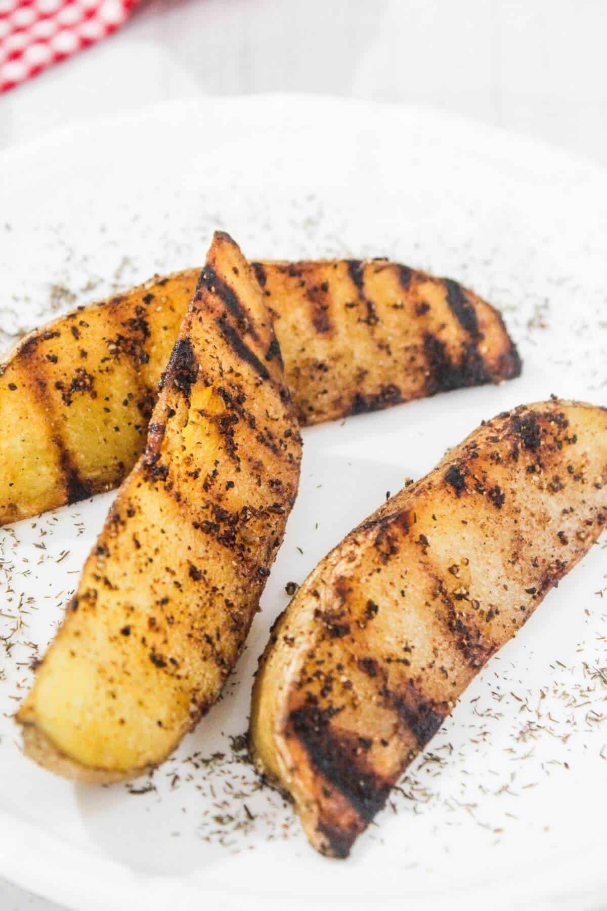 Three grilled potato wedges seasoned with herbs and spices are arranged on a white plate, with scattered dried herbs on the surface.