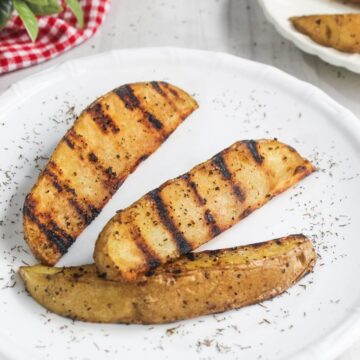Three grilled potato wedges seasoned with herbs and pepper are arranged on a white plate, with dried herbs sprinkled around them.