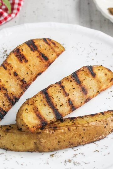 Three grilled potato wedges seasoned with herbs and pepper are arranged on a white plate, with dried herbs sprinkled around them.