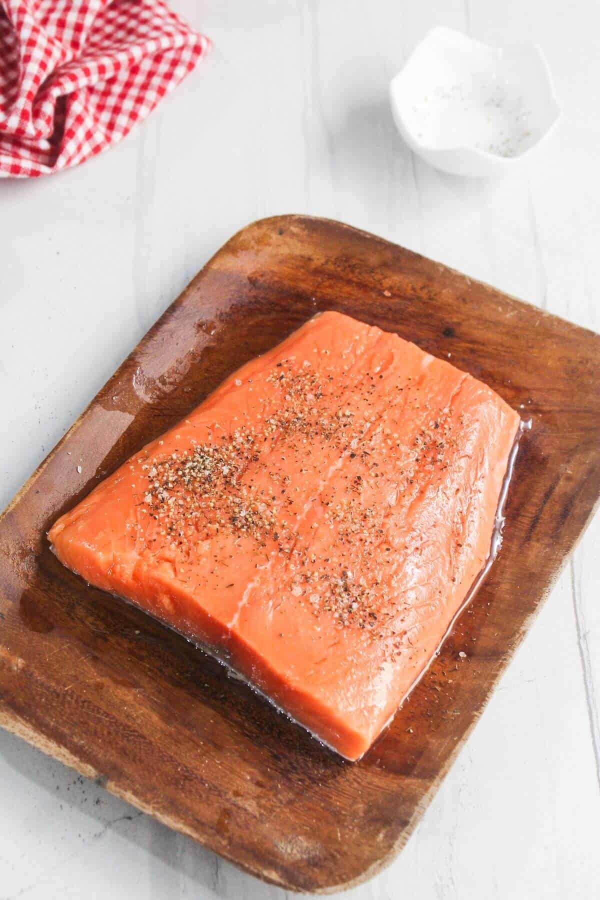 Raw salmon fillet seasoned with salt and pepper on a wooden board, with a red checkered cloth and a bowl of pepper in the background.