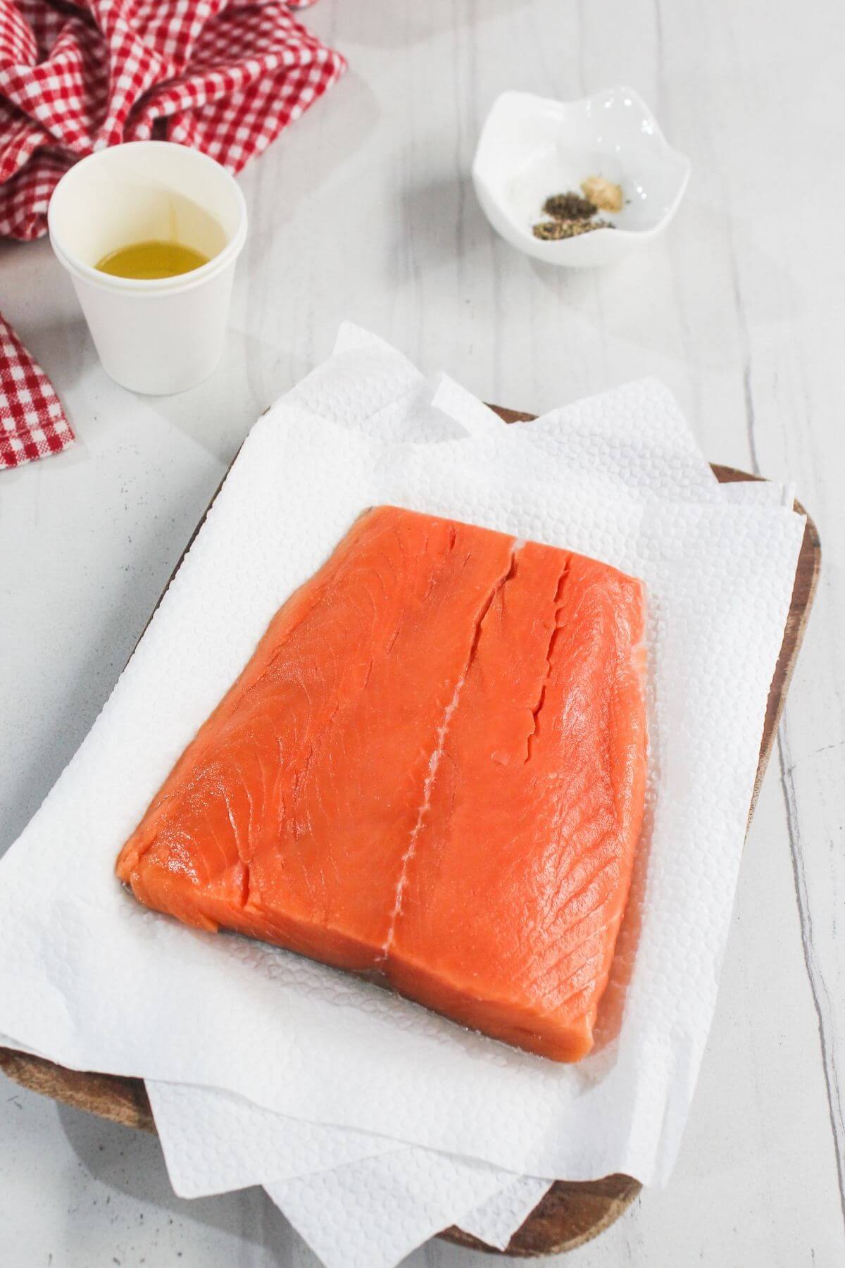 A raw salmon fillet rests on paper towels on a wooden tray, with a cup of oil and a small dish of spices nearby on a white surface.