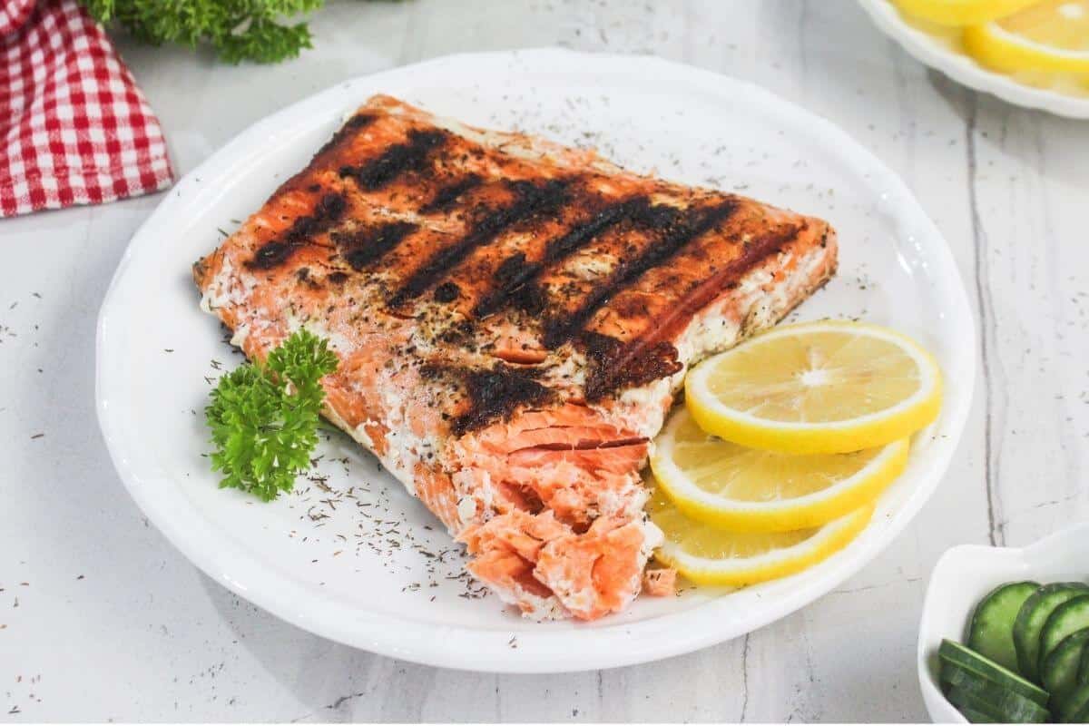 Grilled salmon fillet on a white plate with lemon slices and parsley garnish, sprinkled with herbs, on a white table.