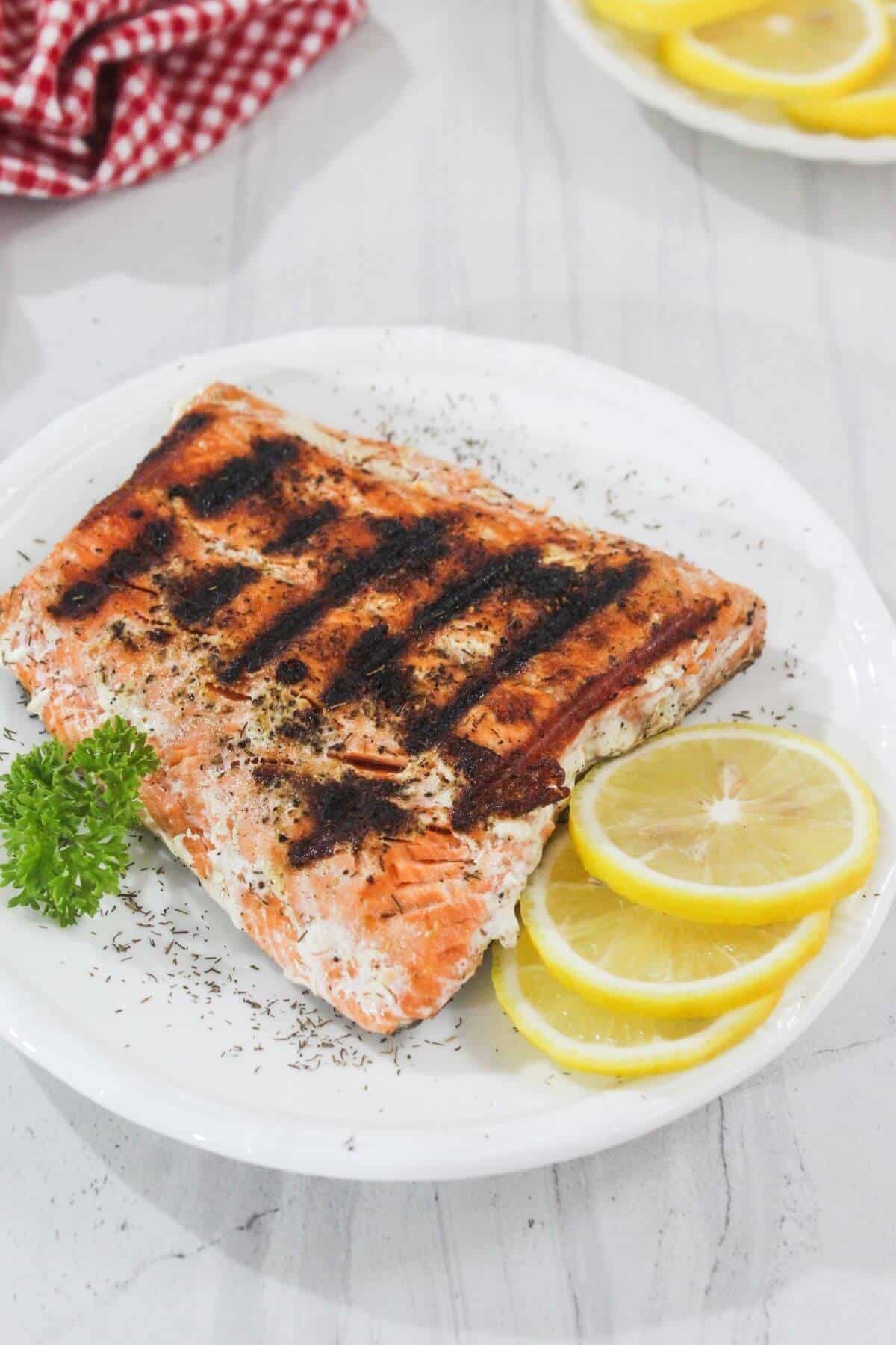 Grilled salmon fillet with char marks on a white plate, garnished with lemon slices, parsley, and dried herbs.