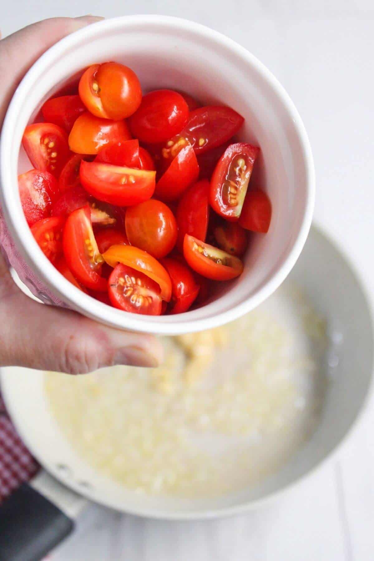 A hand holds a bowl of halved cherry tomatoes over a pan with chopped onions cooking in oil.