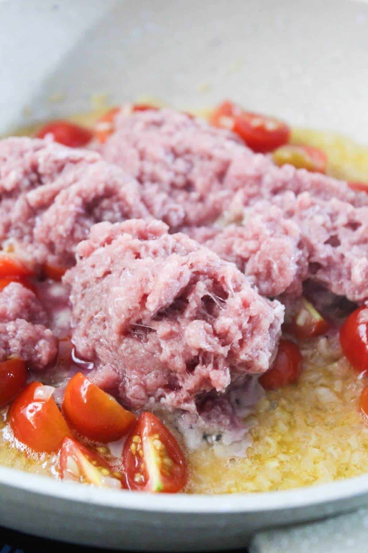 Ground meat and halved cherry tomatoes cooking in a pan with melted butter or oil.