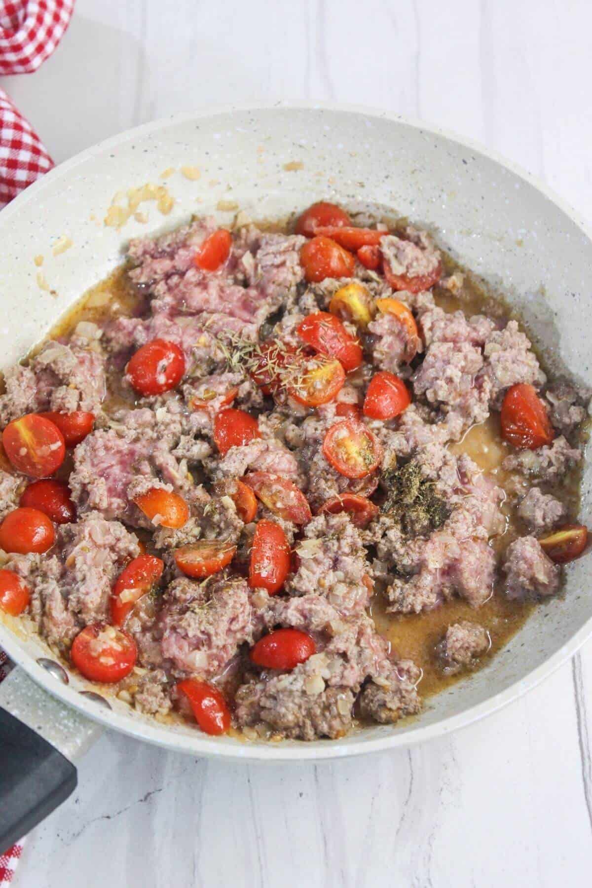 Ground beef cooking in a skillet with halved cherry tomatoes, herbs, and visible juices on a white countertop.