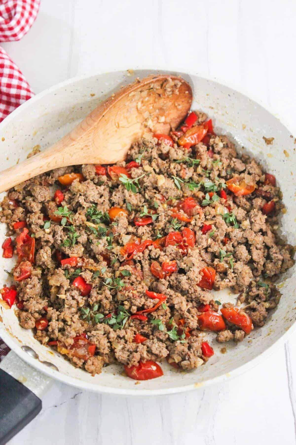 A skillet with cooked ground meat, diced tomatoes, and herbs, stirred with a wooden spoon on a light countertop.