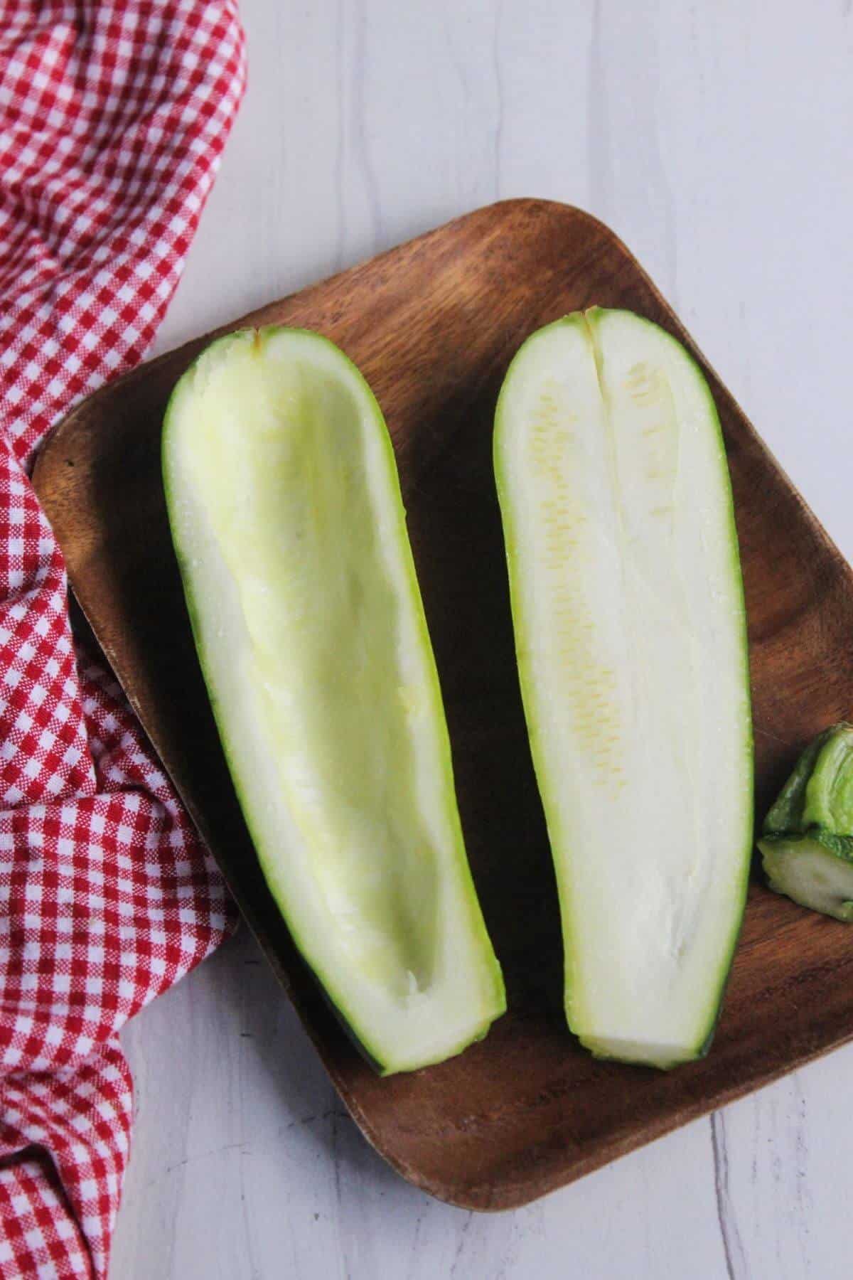 A hollowed-out zucchini cut in half lengthwise sits on a wooden plate next to a red checkered cloth on a white surface.