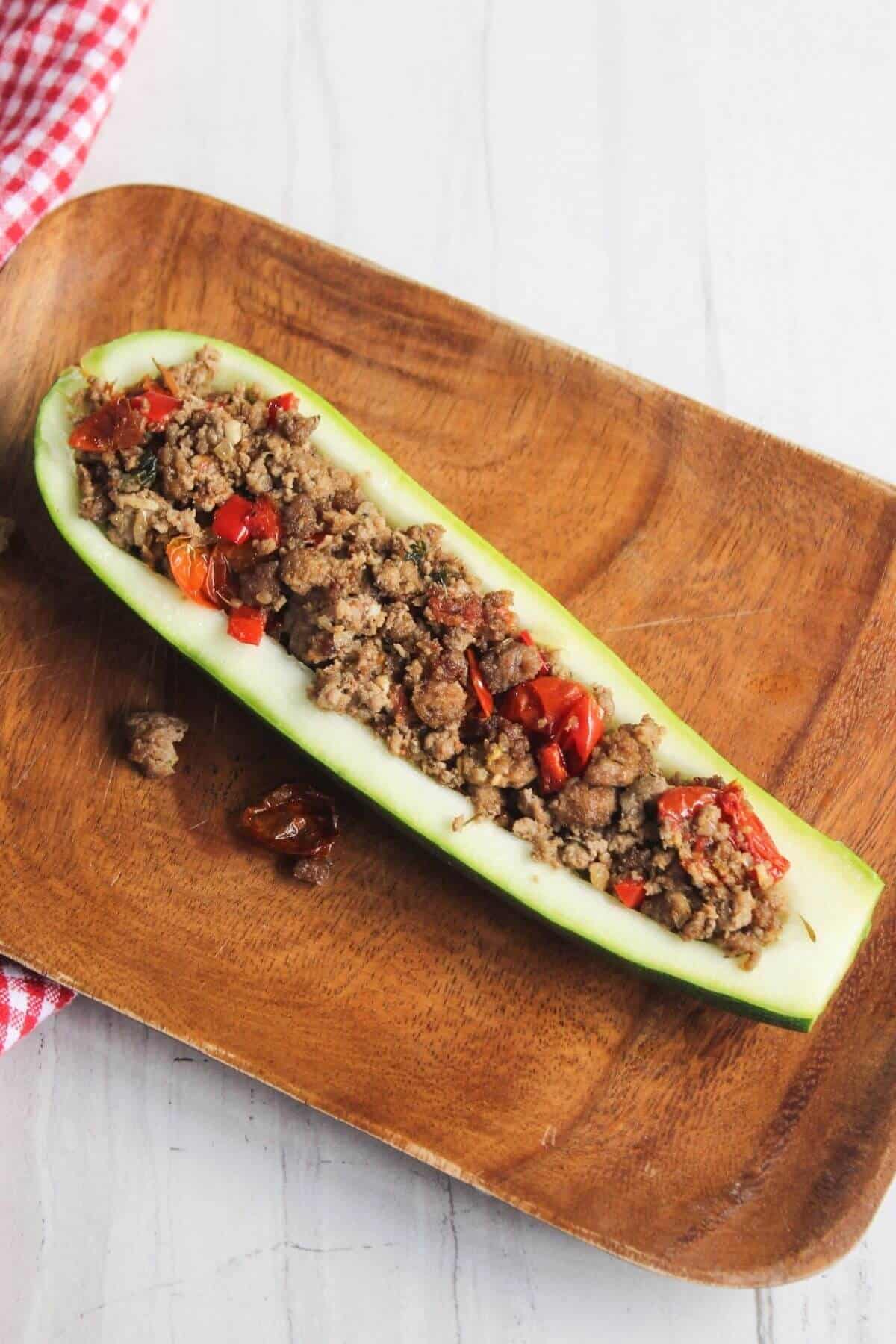 A halved zucchini filled with ground meat and diced red vegetables is served on a wooden plate.