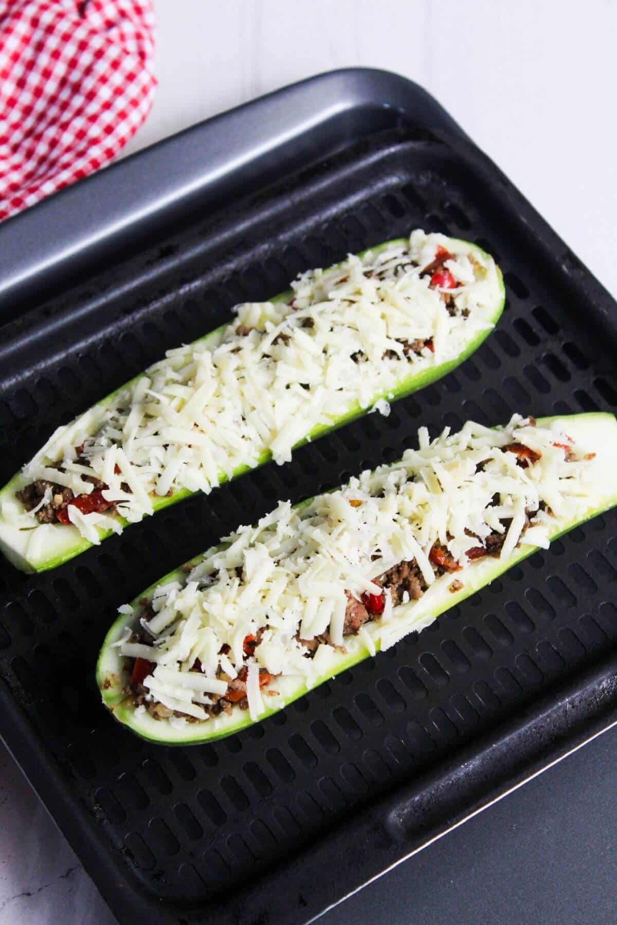 Two zucchini halves stuffed with vegetables and topped with shredded cheese are placed on a black baking tray, ready to be cooked.