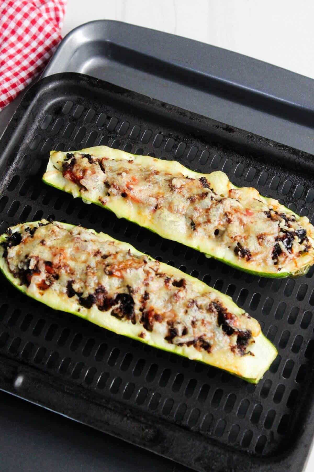 Two zucchini halves filled with a savory mixture and melted cheese are arranged on a black roasting pan.