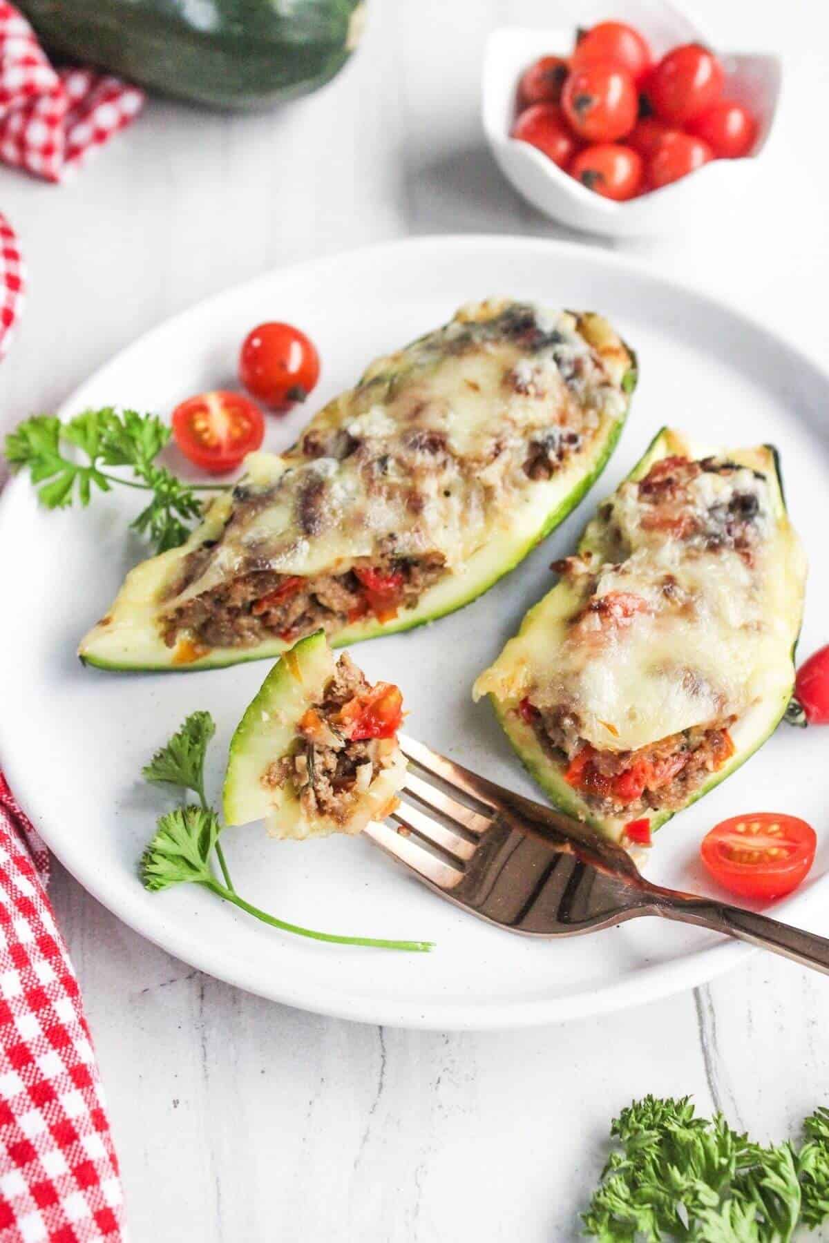 Two zucchini boats stuffed with ground meat, tomatoes, and melted cheese are served on a white plate, garnished with parsley and cherry tomatoes, with a fork holding a small bite.