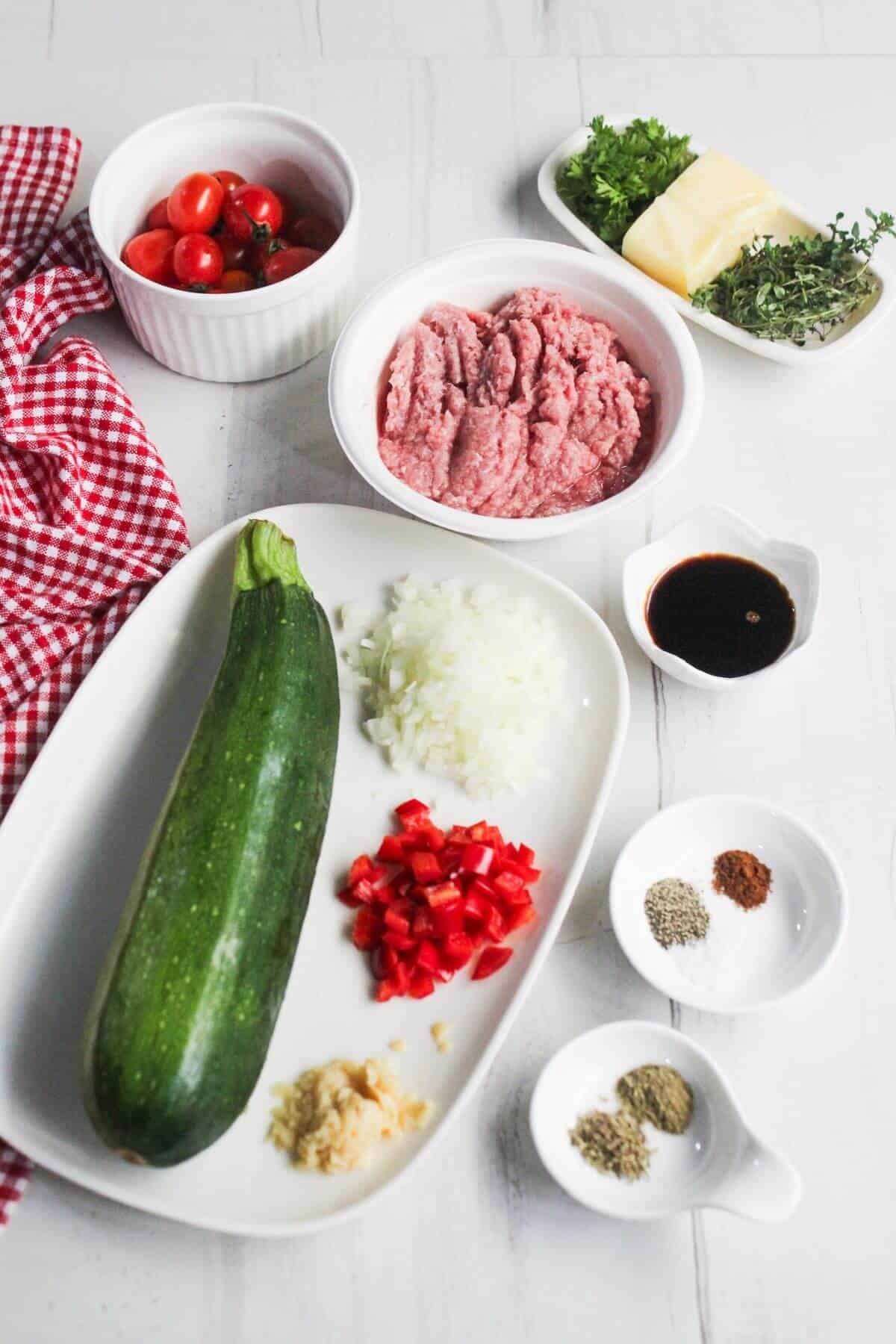 Ingredients arranged on a white surface: zucchini, ground beef, chopped onion, diced red pepper, grape tomatoes, herbs, butter, garlic, spice powders, and a small bowl of dark liquid.