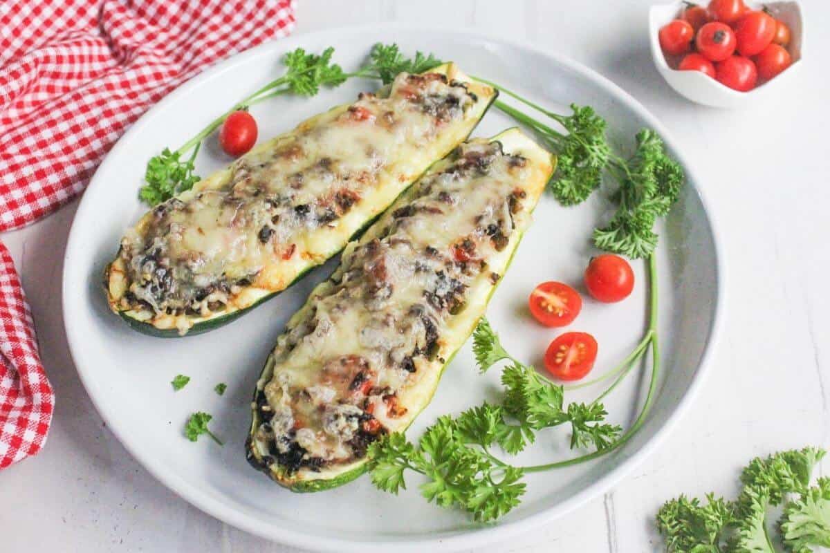 Two zucchini halves stuffed with vegetables and topped with melted cheese are served on a white plate, garnished with parsley and cherry tomatoes. A small bowl of cherry tomatoes is beside the plate.