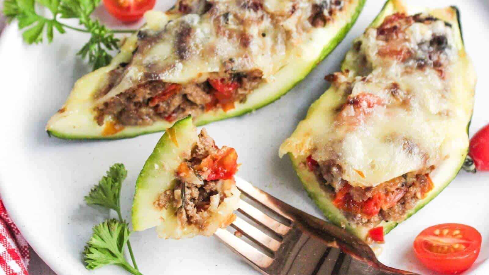 Three zucchini boats filled with ground meat, tomatoes, and melted cheese on a white plate with parsley and tomato garnish; a fork holds a bite-sized piece.
