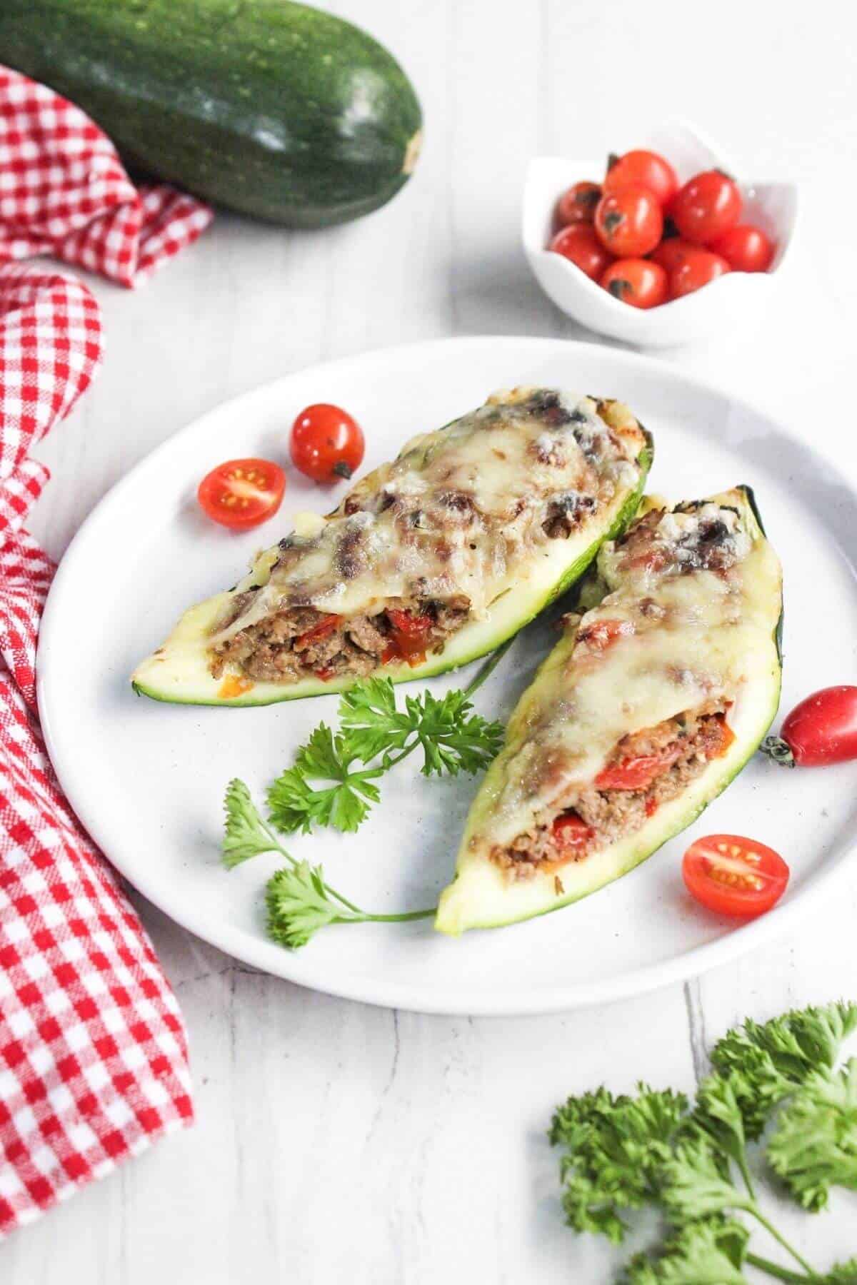 Two zucchini boats filled with ground meat, tomatoes, and melted cheese are served on a white plate with cherry tomatoes and parsley garnish.