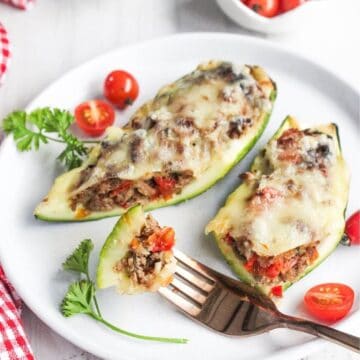 Two zucchini halves stuffed with ground meat, vegetables, and melted cheese on a white plate, garnished with parsley and cherry tomatoes. A fork holds a cut piece.