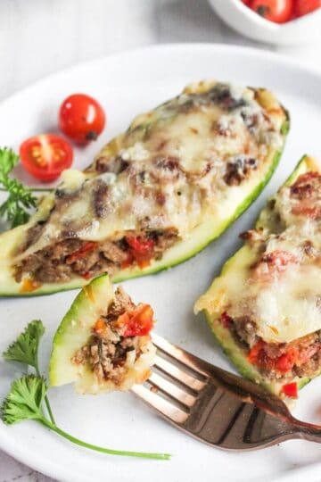 Two zucchini halves stuffed with ground meat, vegetables, and melted cheese on a white plate, garnished with parsley and cherry tomatoes. A fork holds a cut piece.