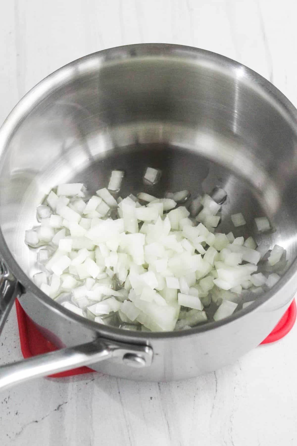 A stainless steel saucepan containing chopped white onions sits on a white surface.