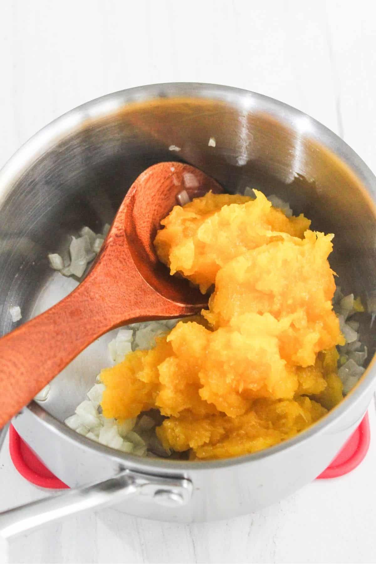 Chopped onions and a scoop of pumpkin puree in a stainless steel saucepan with a wooden spoon on a white surface.