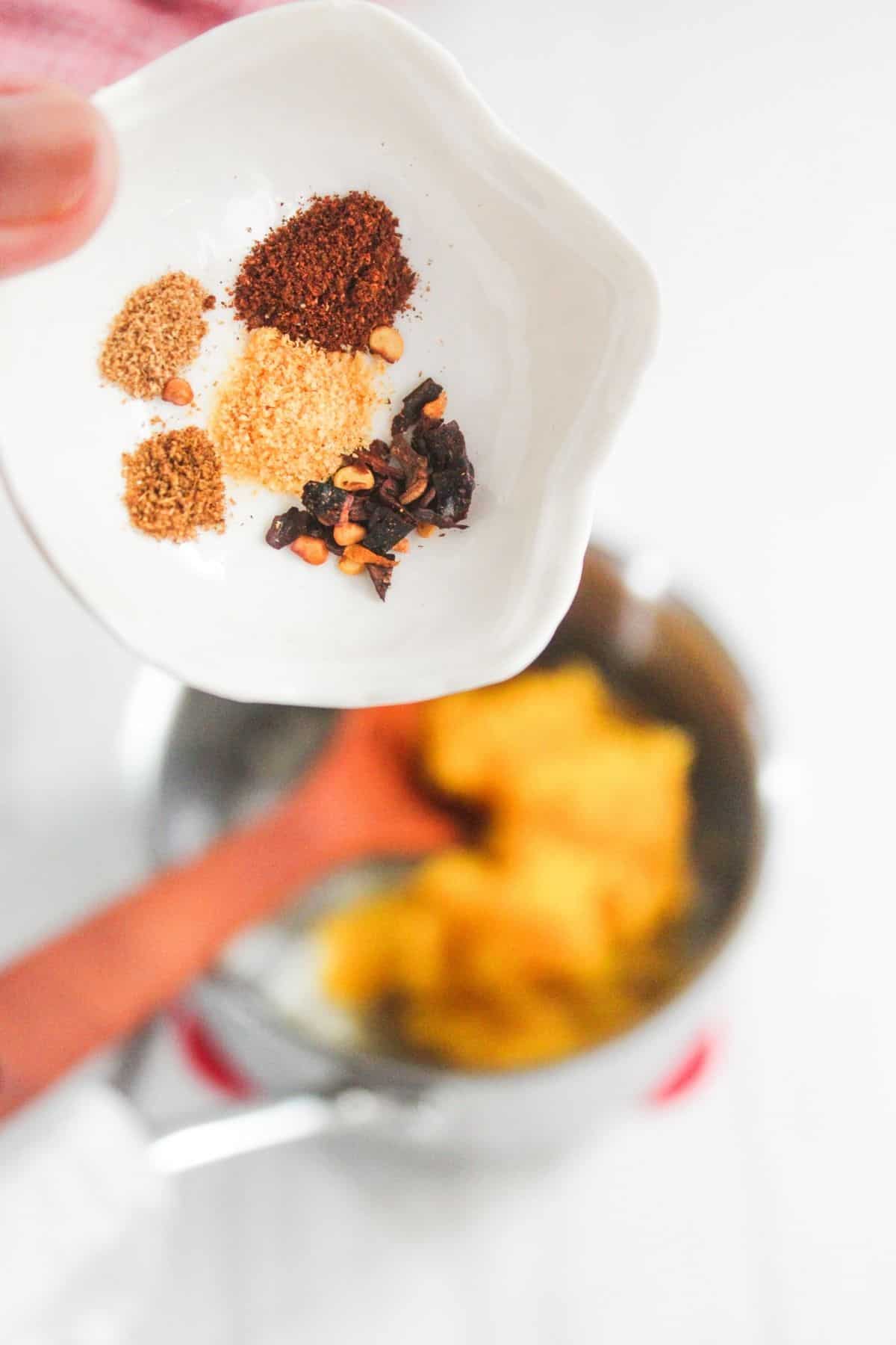 A hand holds a small dish with assorted ground spices above a pot containing yellow ingredients.