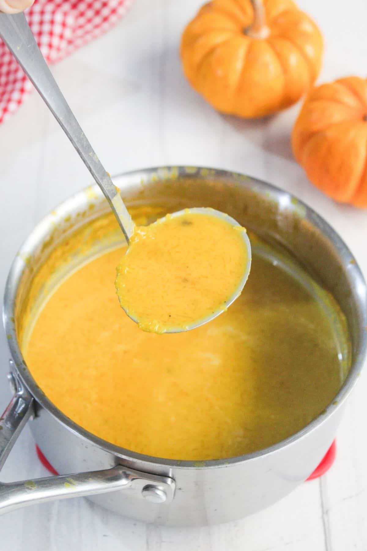 A ladle holds yellow-orange soup above a pot, with two small pumpkins and a red checked cloth in the background.