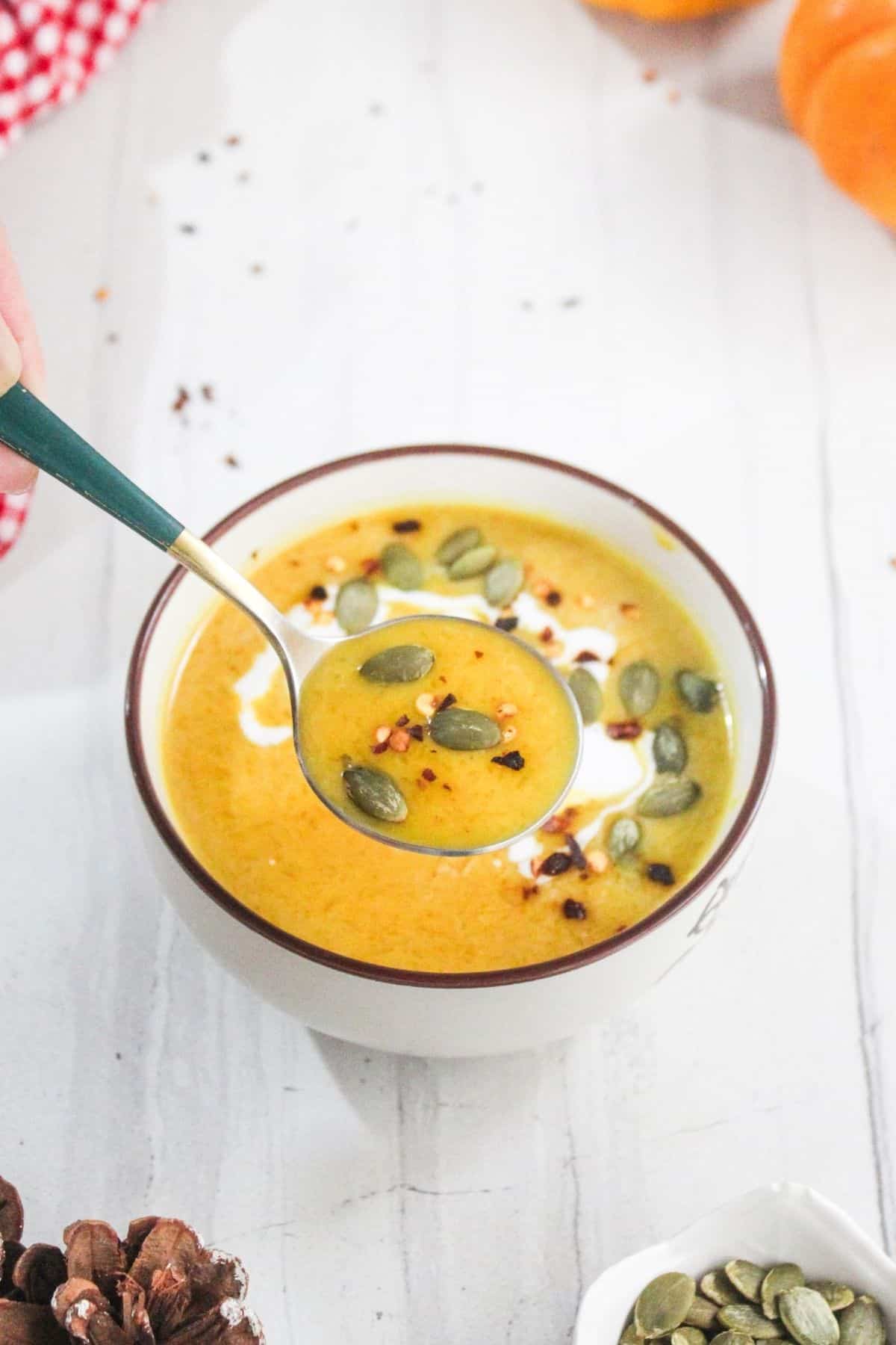 A bowl of creamy orange soup topped with pumpkin seeds and red pepper flakes, with a spoon lifting a portion from the bowl.